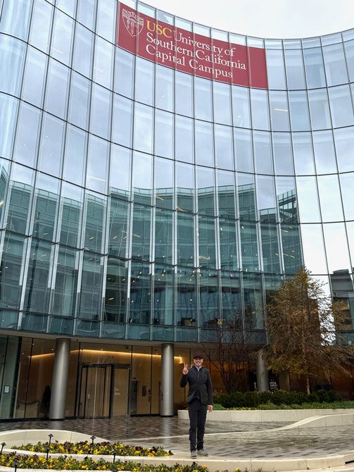 A person stands in front of a modern, curved glass building with a red sign reading "USC University of Southern California Capital Campus," holding up a peace sign.