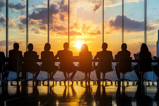 2025 Board of Directors and Staff; A group of people in suits sit at a conference table, silhouetted against a large window. A vibrant sunset with orange and pink hues is visible outside.