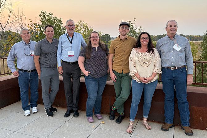 2025 Board of Directors and Staff; A group of seven people standing outdoors on a patio with a sunset in the background. They are smiling and dressed casually, conveying a relaxed atmosphere.