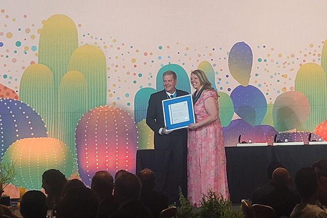 A man and a woman smile while holding a framed certificate against a colorful abstract backdrop. The scene is warm, celebratory, and formal.