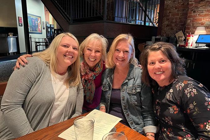 Four women sit closely together at a restaurant table, smiling warmly. They appear joyful and relaxed, suggesting a friendly gathering.