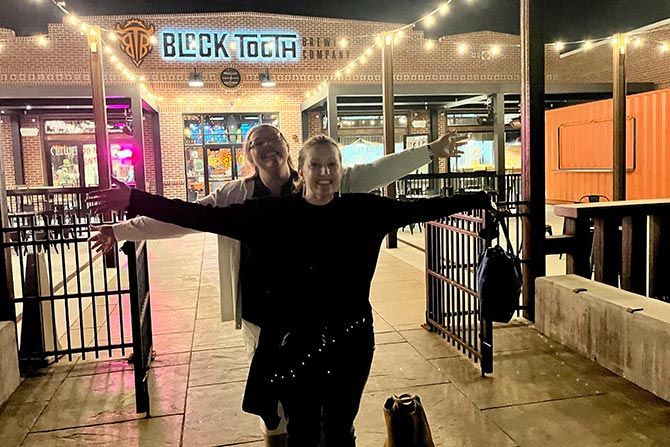 Two people happily pose with arms outstretched in front of a brightly lit building at night. The sign above reads "Black Tooth Brewing Company."