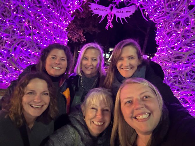 Six smiling women pose in front of a large, illuminated archway made of antlers adorned with purple lights. The atmosphere is joyful and festive.