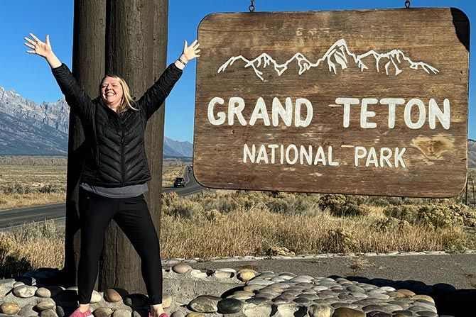A Tribute to Emily Cronbaugh Bogstie; A joyful woman with outstretched arms stands beside the Grand Teton National Park sign. Mountains and clear skies form a scenic backdrop.