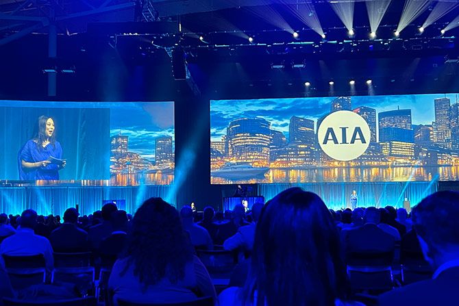 AIA Conference on Architecture & Design 2025; Audience facing a large stage with illuminated screens displaying a city skyline and "AIA." A presenter stands on stage, creating an engaging atmosphere.