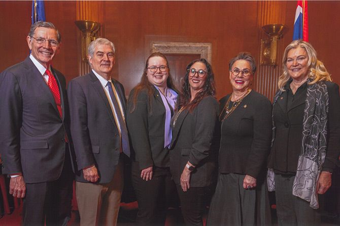 AIA Leadership Summit 2025; A group of six adults dressed in formal attire stands in a room with wooden paneling. They all smile warmly, conveying a sense of camaraderie.