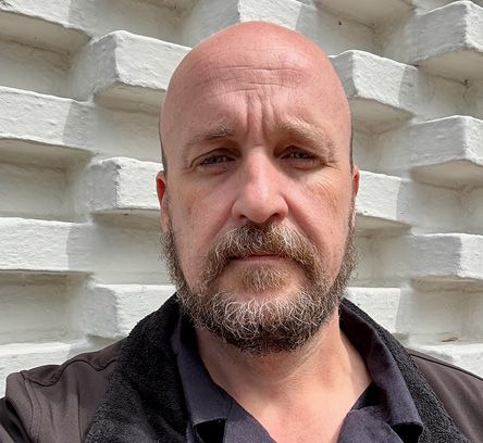 Jeff Huber; A bald man with a beard stands in front of a textured white brick wall. He's wearing a black shirt and jacket, looking directly at the camera with a neutral expression.