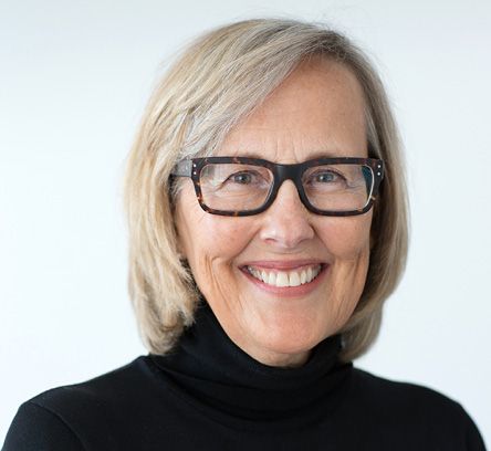Julie Snow; A smiling older woman with glasses and blonde hair, wearing a black turtleneck. She stands against a plain white background, conveying warmth and friendliness.
