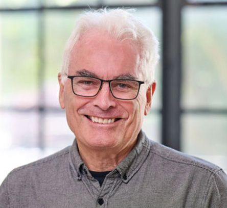 William Leddy; Elderly man with white hair and glasses smiling warmly, wearing a gray shirt. Background features softly blurred windows suggesting a bright setting.