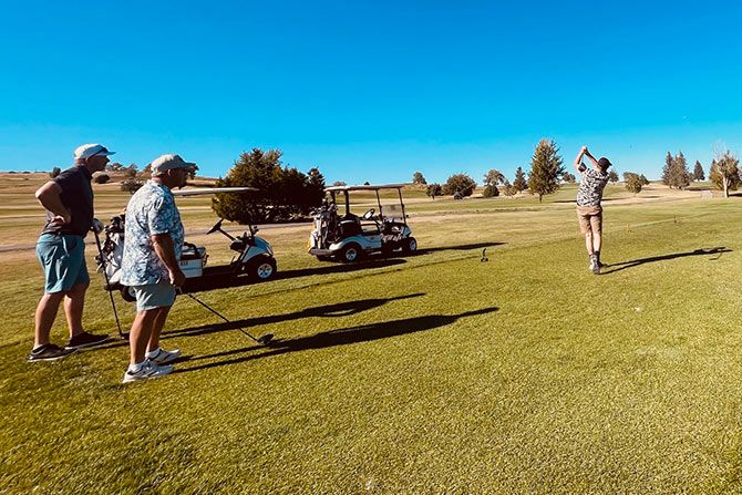 Architectural Education Foundation; A golfer swings on a sunlit course, watched by others with clubs. Golf carts and trees are in the background. The mood is relaxed and focused.