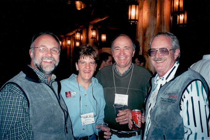 Four people stand closely together at a dimly lit indoor event, smiling warmly. They wear casual outfits with name tags and hold drinks, suggesting a relaxed, friendly gathering.