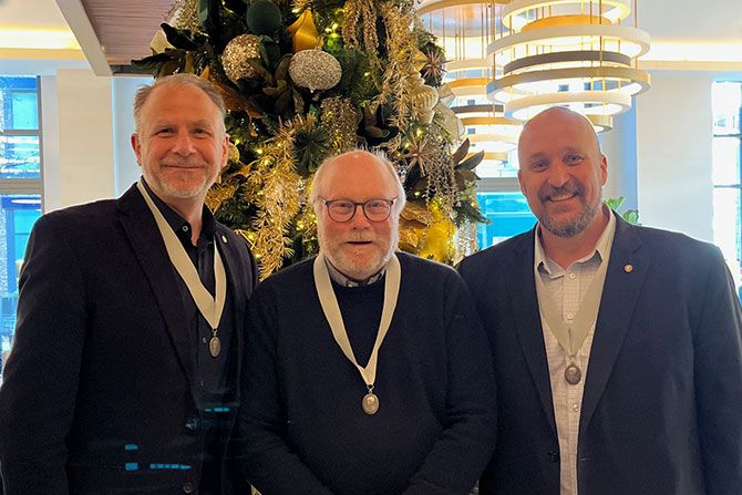 Individual Honors: Dan Stalker, AIA; Three men smiling and wearing medals stand in front of a decorated Christmas tree. The setting is bright and festive, conveying a joyful atmosphere.
