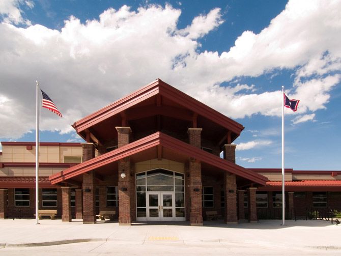 Front view of a modern brick building with a red gabled roof, flanked by two flagpoles. A bright blue sky and fluffy clouds are overhead.