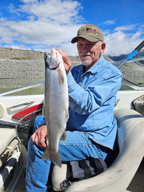 An older man in a denim shirt and cap sits in a boat holding a large fish he caught, with a mountainous landscape and blue sky in the background.