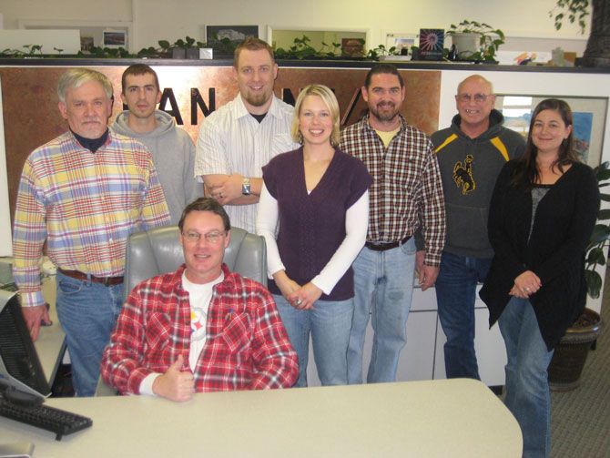 A group of eight people, seven standing and one seated, smiling in an office. The seated person gives a thumbs-up. The mood is friendly and casual.