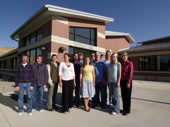 A group of ten people stands smiling in front of a modern brick building under a clear blue sky. The building has a "Welcome" sign above the entrance.