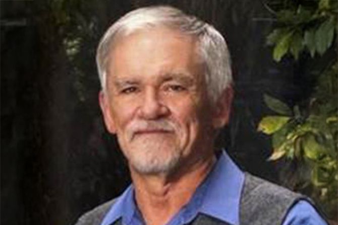 In Memoriam: David Gerald Wellington; An elderly man with white hair and a beard, wearing a blue shirt and gray vest, smiles gently against a backdrop of greenery, conveying warmth and wisdom.