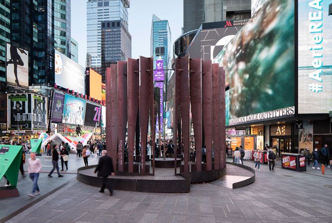 Urban installation in Times Square features vertical brown panels forming a circular structure. Vibrant billboards and blurred crowds surround the scene.