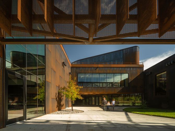 Modern building with rust-colored metal facade and large windows, featuring landscaped courtyard. Two people walk, conveying a calm and serene atmosphere.