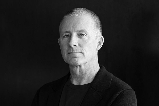 Eric Logan; Black and white portrait of an older man with a calm expression, wearing a dark jacket and shirt, against a dark background. Professional and contemplative tone.