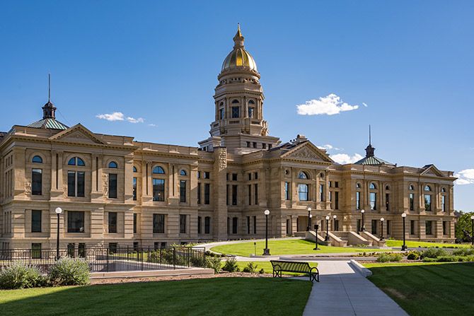 Government Advocacy Committee; Historic building with a golden dome and detailed stone architecture under a clear blue sky. Lush greenery and a bench adorn the foreground.