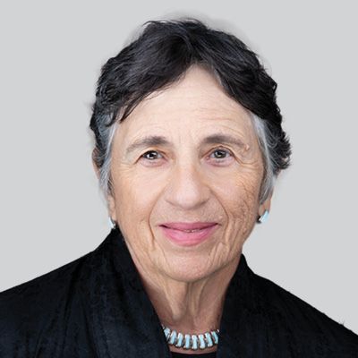 Carol Ross Barney; Elderly woman with short gray hair and kind expression, wearing a black jacket and turquoise necklace, set against a plain gray background.