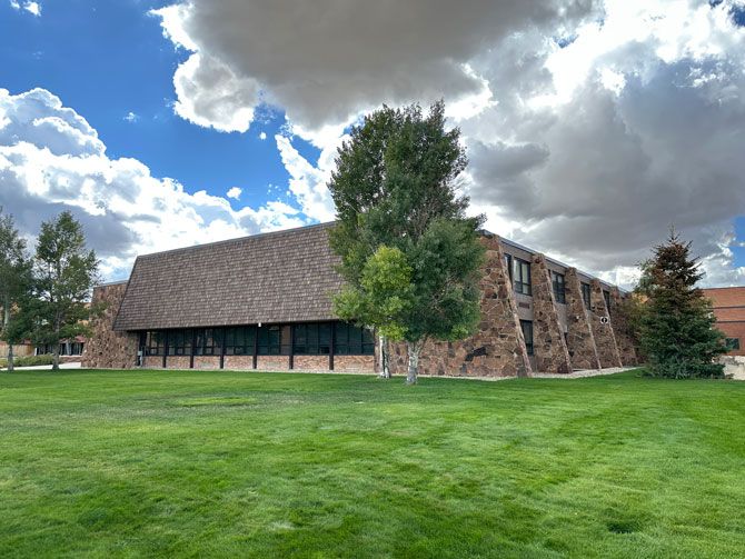 A large building with sloped brown shingle roof and stone walls stands against a blue sky with fluffy clouds. Green grass and trees are in the foreground.