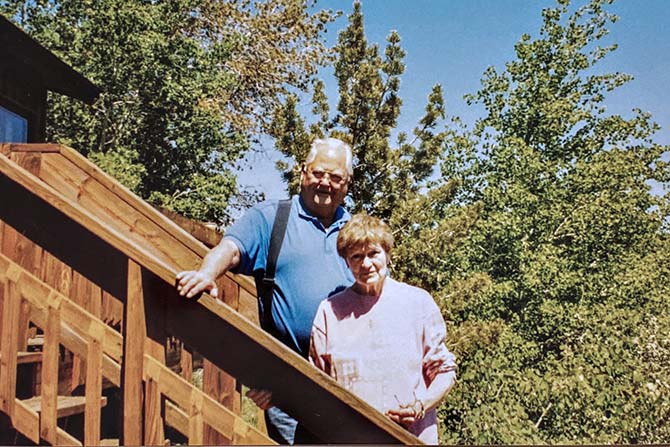 A smiling elderly couple stands on a wooden outdoor staircase surrounded by lush greenery and clear blue skies, conveying a relaxed, joyful atmosphere.