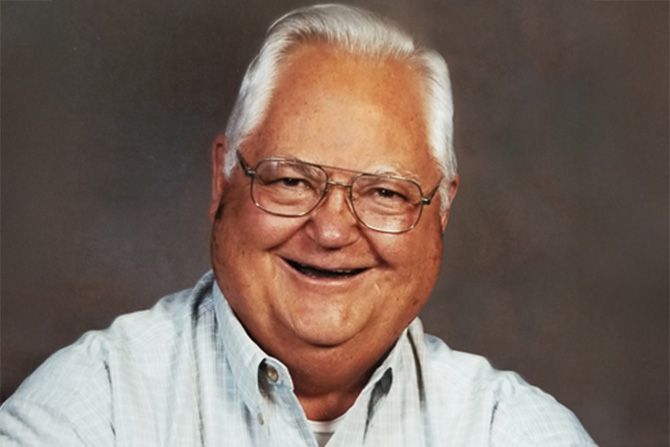 John L. Frullo; a tan man with white hair and glasses smiling with a dark brown background.