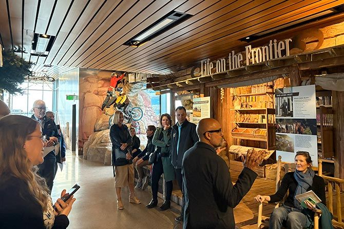 Membership Development and Education & Resources Committees; A group of people stand in a museum exhibit titled "Life on the Frontier," featuring a rustic interior and a motorcycle on display. The atmosphere is engaging.