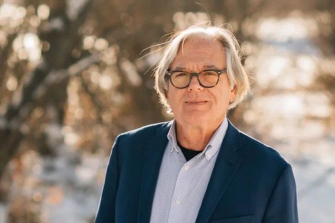A Conversation With Tom Ward; Elderly man with glasses, wearing a blue suit and white shirt, stands outdoors with sunlight filtering through trees. The atmosphere is serene and warm.